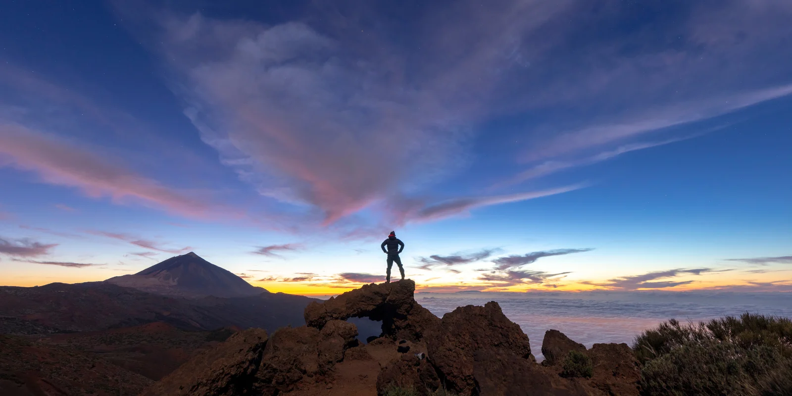 Paisaje volcánico al atardecer en Tenerife con el Teide de fondo y una persona con los brazos en la cintura mirando hacia el horizonte