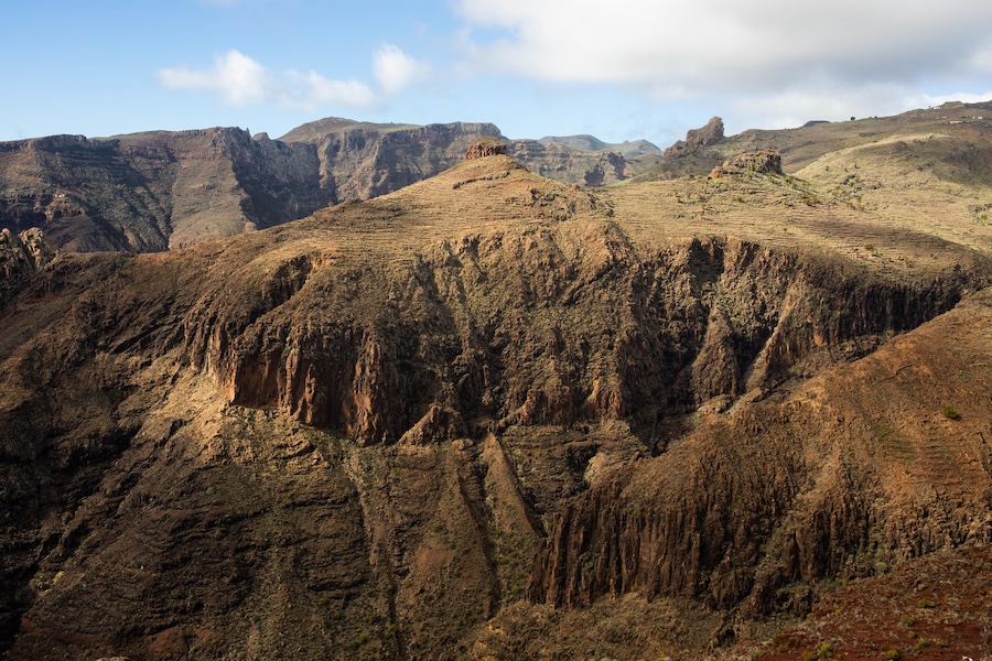 Altos acantilados volcánicos de La Gomera cayendo al océano Atlántico con olas rompiendo en la base y luz cálida al atardecer.